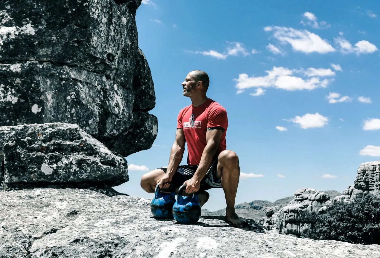 A muscular man exercises with kettlebells on rocky ground under a bright blue sky, emphasizing strength and fitness.